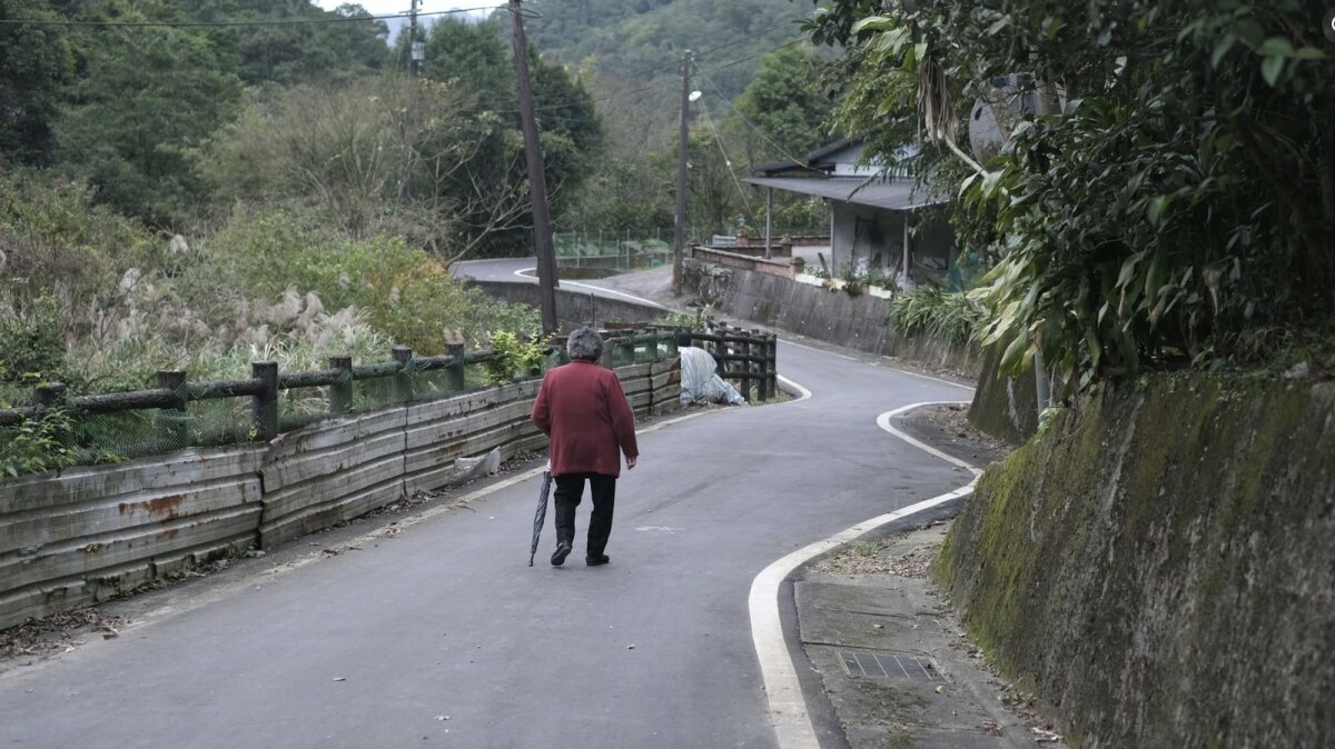 一位長者拄著拐杖走在彎曲的山區道路上，沿途有護欄與樹木